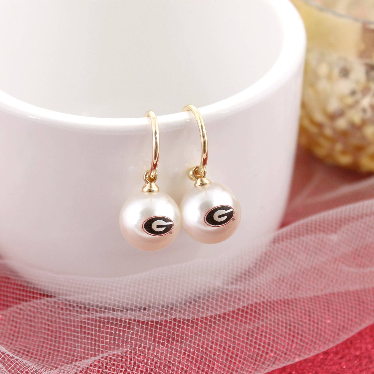 Gold hoop earrings with white pearls featuring Georgia “G” logo, displayed on a white background with red fabric accent.