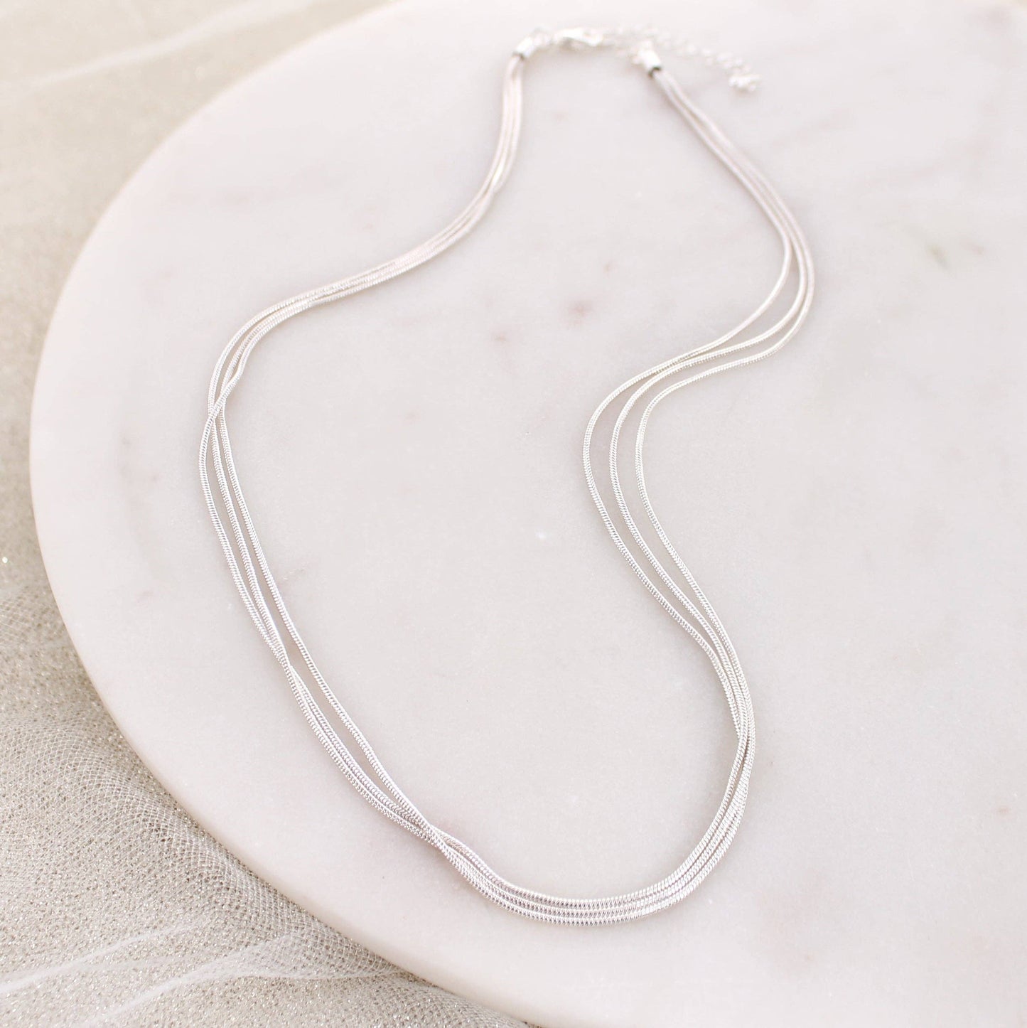 Silver three-strand snake chain necklace displayed on a white marble background.