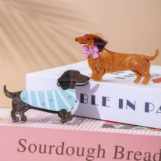 Two dachshund-shaped hair claws, one with a pink bow and one with a blue striped outfit, on a glossy base.