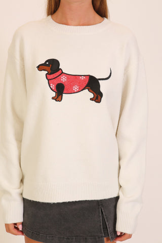 A woman wearing a cream crew-neck pullover sweater with a graphic of a dachshund in a red snowflake-print coat centered on the chest, paired with a dark gray mini skirt.