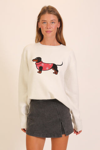 A woman wearing a cream crew-neck pullover sweater with a graphic of a dachshund in a red snowflake-print coat centered on the chest, paired with a dark gray mini skirt.