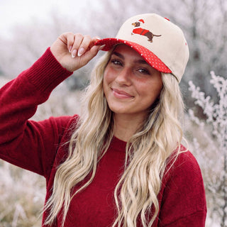 Beige trucker hat with a dachshund wearing a Santa hat, modeled by a woman with long blonde hair.