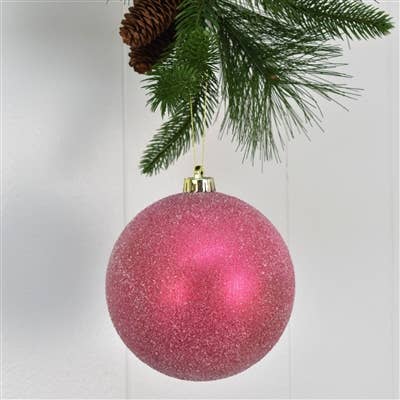 Pink glittery Christmas ball ornament hanging on a tree branch with a pine cone.