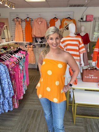 A woman modeling a bright orange strapless top with large white polka dots, paired with blue jeans, inside a clothing boutique. The store features racks of colorful garments, mannequins, and a sign that reads 'THE TOT BAG'.