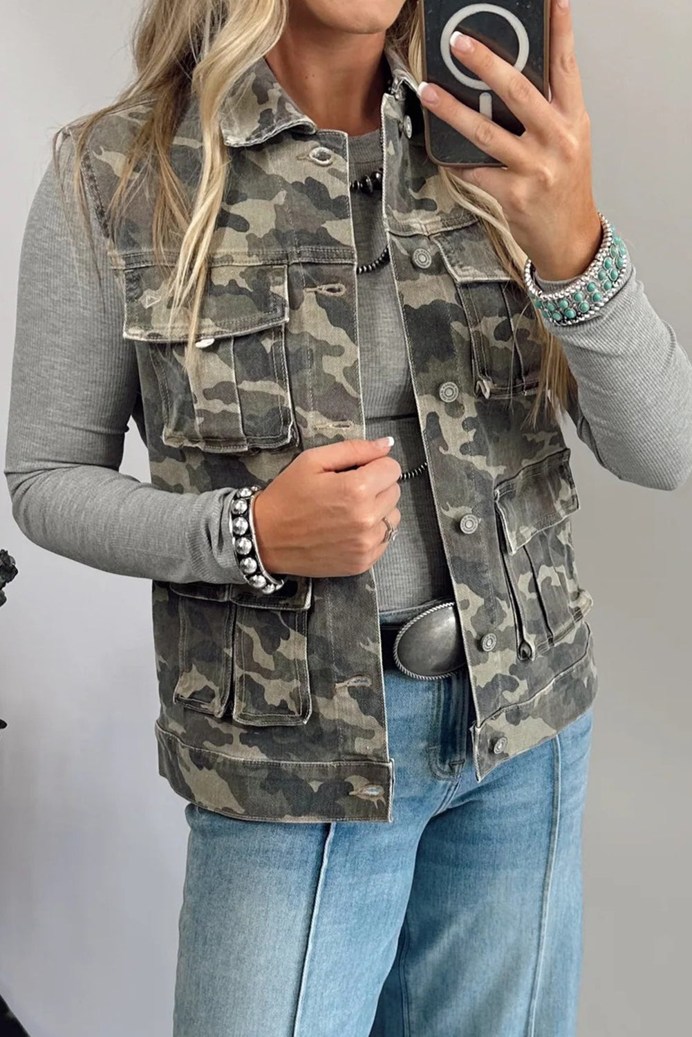 Woman wearing a camo cargo denim vest layered over a gray long-sleeve top and jeans.