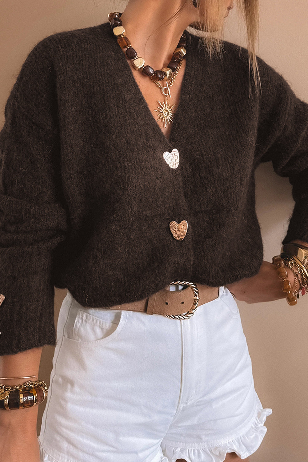 Model wearing a coffee-colored V-neck cardigan with metal heart buttons paired with white shorts and layered jewelry.