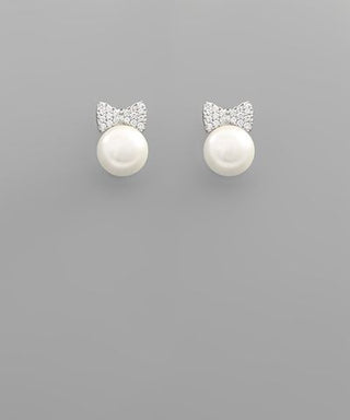 Pearl stud earrings with crystal ribbon bow detail in gold and rhodium finishes displayed on a gray background from The Pink Doxie Boutique.