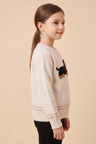A girl wearing a cream fine gauge knit crew neck pullover sweater with a textured black and tan dachshund appliqué on the front and ribbed cuffs and hem.