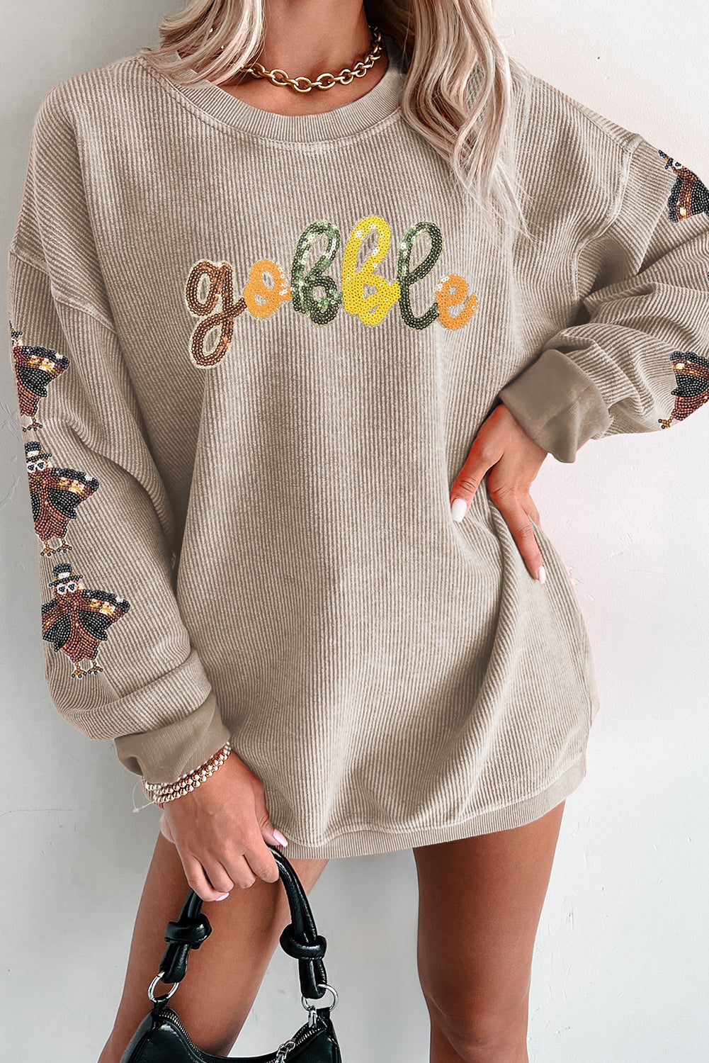 Woman wearing a beige ribbed sweatshirt with colorful sequin “gobble” letters and turkey sequin sleeve patches — festive Thanksgiving top from The Pink Doxie Boutique