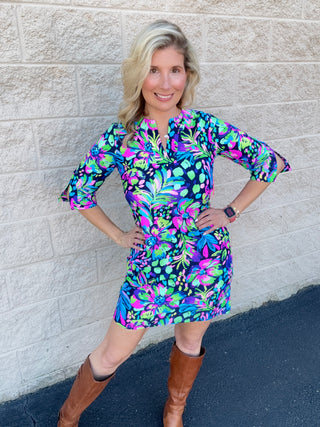 A woman wearing a navy shift dress with a bold multicolor tropical floral print in fuchsia, lime green, turquoise, and purple, featuring a split V-neckline and three-quarter sleeves, styled with tall cognac riding boots.