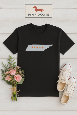 Black JESUS Tennessee Faith Tee displayed flat on a white wood background with flowers and sneakers, Southern preppy boutique style.