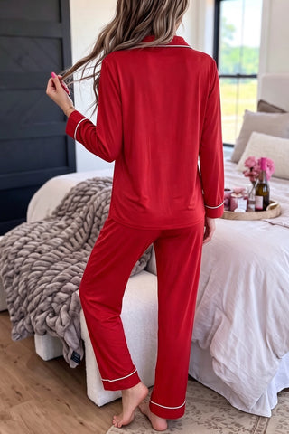 A woman wearing a red two-piece long-sleeve pajama set with white contrast piping trim, a bow embroidery on the chest pocket, and full-length straight-leg drawstring pants.