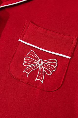 A woman wearing a red two-piece long-sleeve pajama set with white contrast piping trim, a bow embroidery on the chest pocket, and full-length straight-leg drawstring pants.