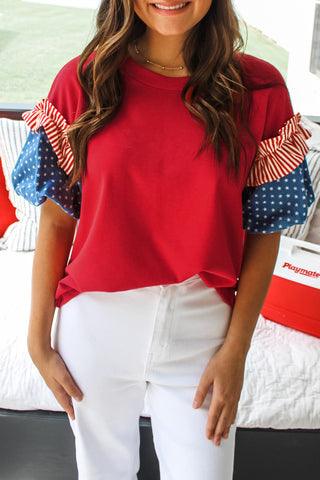 Red stars and stripes puff sleeve top with crew neckline and patriotic details, styled with white pants from The Pink Doxie Boutique.