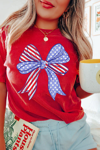 Red patriotic graphic T-shirt with large American flag bow print, styled casually for Fourth of July from The Pink Doxie Boutique.