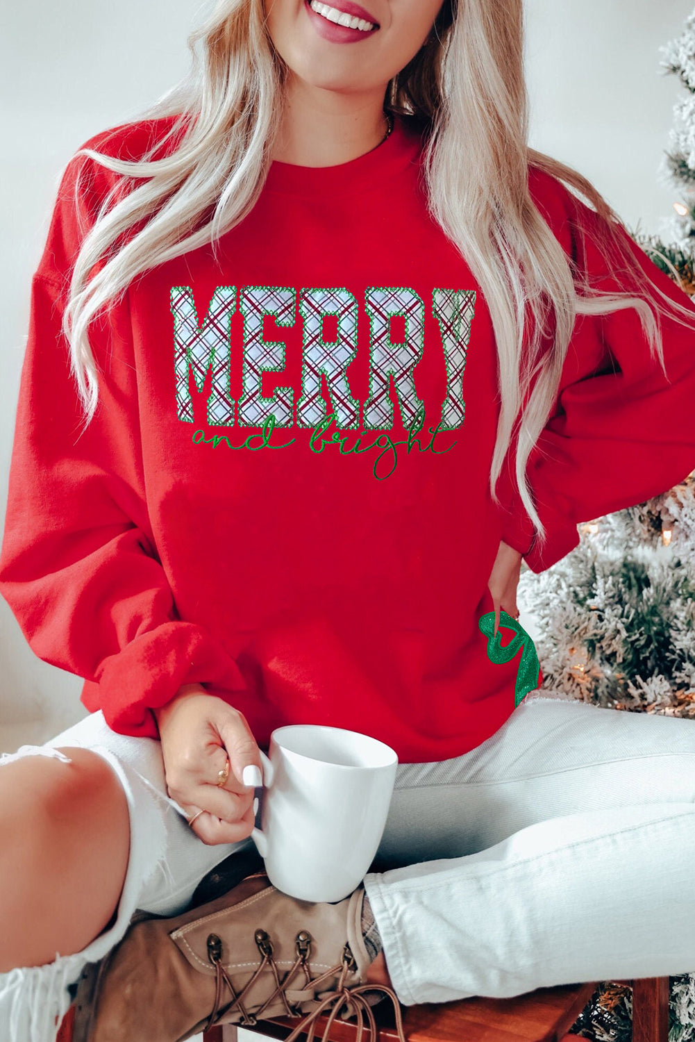 Red Christmas sweatshirt with plaid “MERRY and bright” embroidery and a green sequin bow, worn by a woman sitting with a mug near a Christmas tree.