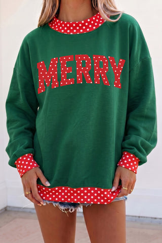 An oversized forest green pullover sweatshirt with a red and white polka dot crew neckline, matching cuffs and hem trim, and a bold arched "MERRY" graphic on the chest in red polka dot lettering.