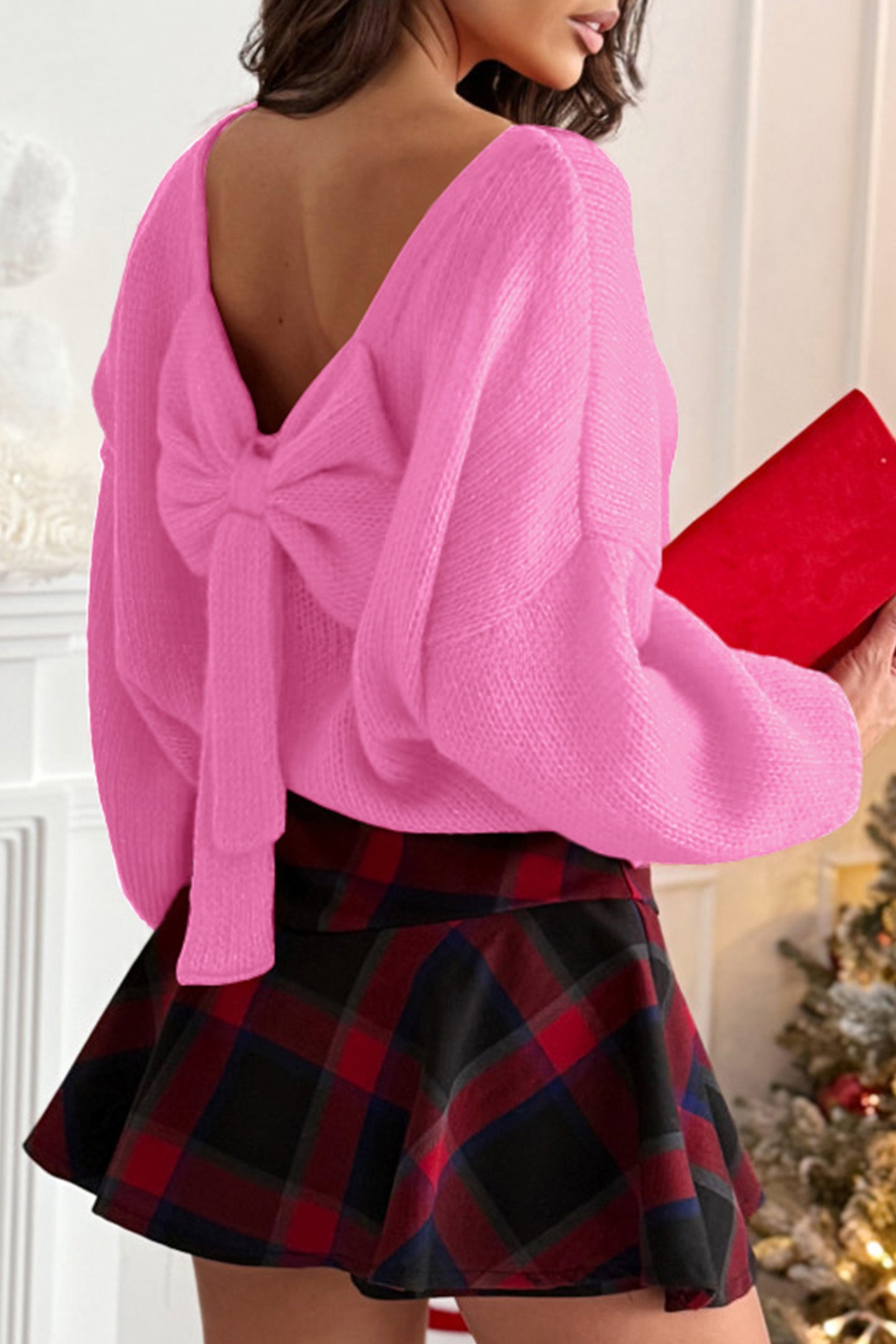 Woman wearing a pink open-back sweater with an oversized bow and bubble sleeves, styled with a plaid mini skirt from The Pink Doxie Boutique.