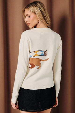 A woman wearing a cream button-down collared cardigan with a large knit dachshund graphic wearing an argyle scarf in blue, yellow, and brown, paired with a dark denim mini skirt.