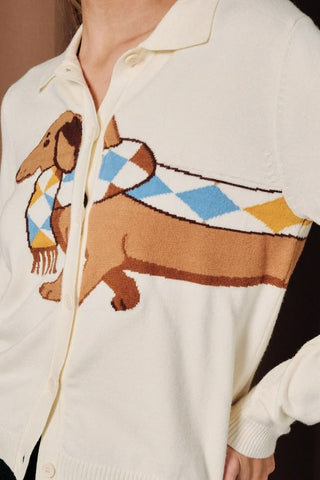 A woman wearing a cream button-down collared cardigan with a large knit dachshund graphic wearing an argyle scarf in blue, yellow, and brown, paired with a dark denim mini skirt.