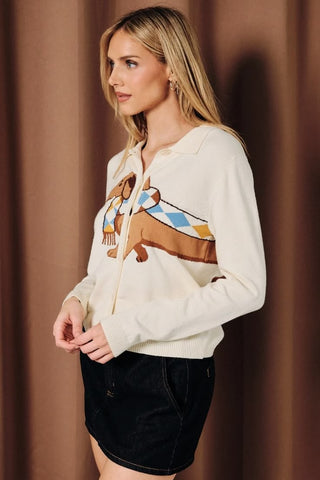 A woman wearing a cream button-down collared cardigan with a large knit dachshund graphic wearing an argyle scarf in blue, yellow, and brown, paired with a dark denim mini skirt.