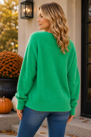 A woman wearing a relaxed-fit, fuzzy kelly green button-front cardigan with a v-neckline and long sleeves, paired with blue jeans.