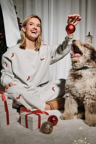 A woman wearing a relaxed-fit heather gray crew neck sweatshirt covered in small embroidered dachshunds dressed in red holiday outfits, styled with matching gray jogger pants.