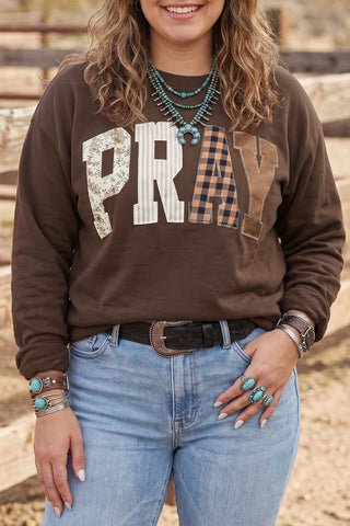 A plus-size woman wearing a chocolate brown pullover sweatshirt with large patchwork appliqué letters spelling PRAY across the chest, each letter in a different fabric pattern including floral, stripe, buffalo check, and solid, paired with light wash jeans and turquoise jewelry.