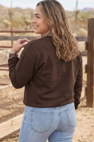 A plus-size woman wearing a chocolate brown pullover sweatshirt with large patchwork appliqué letters spelling PRAY across the chest, each letter in a different fabric pattern including floral, stripe, buffalo check, and solid, paired with light wash jeans and turquoise jewelry.