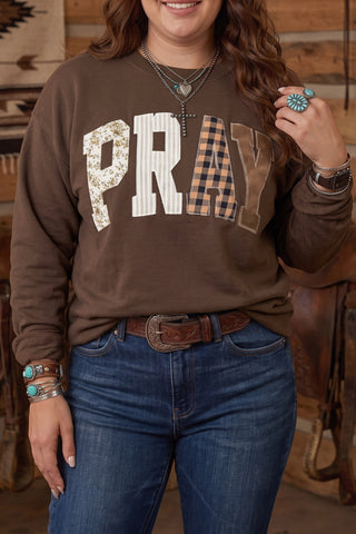 A plus-size woman wearing a chocolate brown pullover sweatshirt with large patchwork appliqué letters spelling PRAY across the chest, each letter in a different fabric pattern including floral, stripe, buffalo check, and solid, paired with light wash jeans and turquoise jewelry.