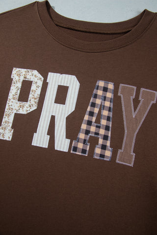 A plus-size woman wearing a chocolate brown pullover sweatshirt with large patchwork appliqué letters spelling PRAY across the chest, each letter in a different fabric pattern including floral, stripe, buffalo check, and solid, paired with light wash jeans and turquoise jewelry.