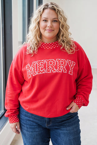 A plus-size model wearing a red pullover sweatshirt with a varsity-style polka dot "MERRY" graphic on the chest, a red-and-white polka dot mock-neck collar, and matching polka dot cuff trim, paired with dark-wash jeans.