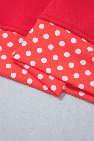 A plus-size model wearing a red pullover sweatshirt with a varsity-style polka dot "MERRY" graphic on the chest, a red-and-white polka dot mock-neck collar, and matching polka dot cuff trim, paired with dark-wash jeans.