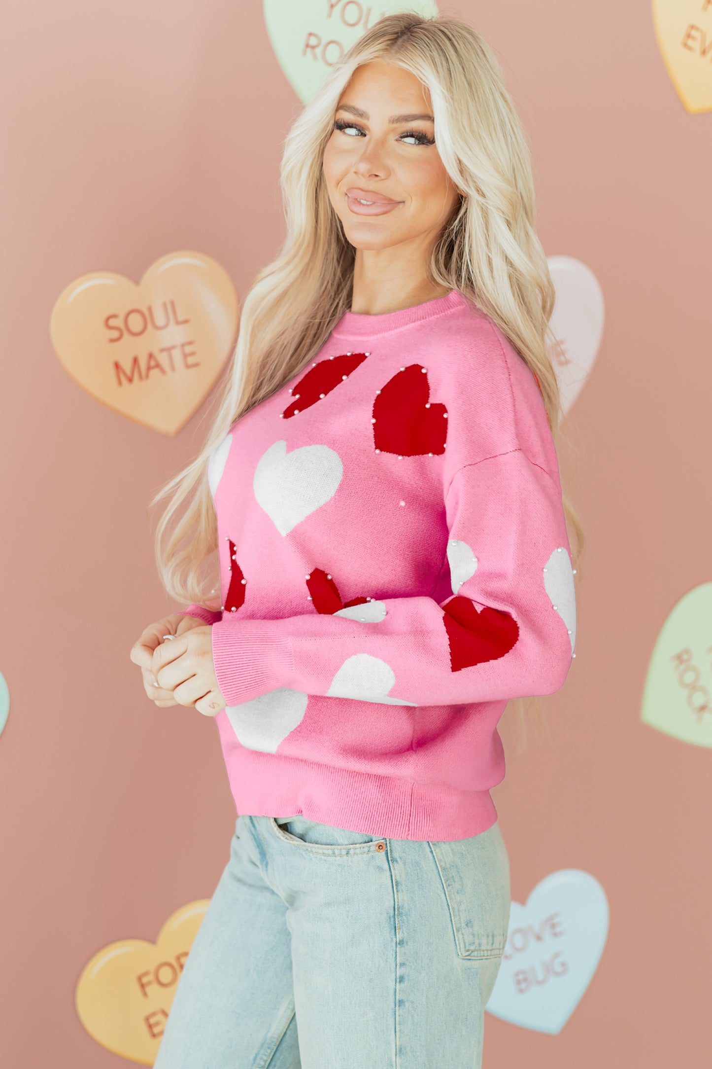 Woman wearing a pink Valentine’s sweater with pearl-embellished red and white hearts, posing against a pastel heart-themed backdrop.