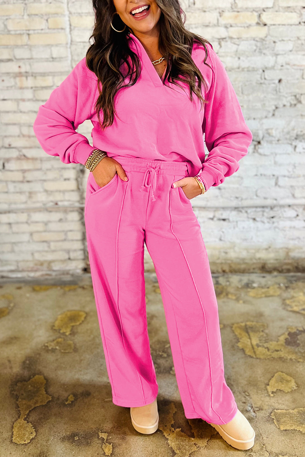 Woman wearing a bright pink two-piece set with a collared V-neck pullover and wide-leg seam pants, standing against a light brick wall background.