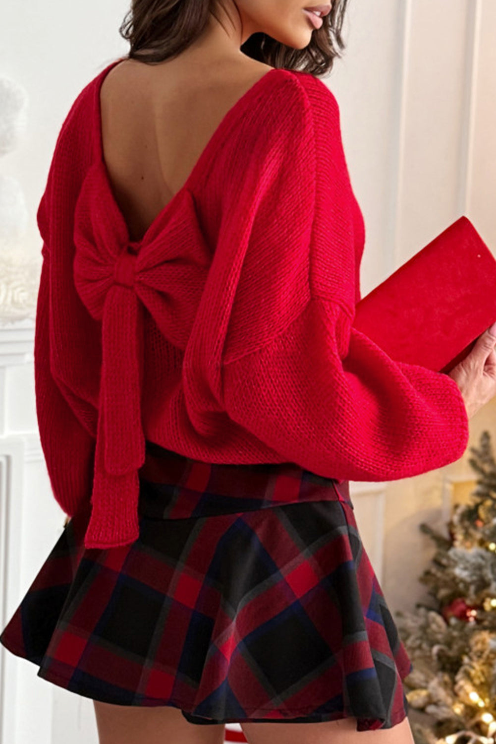 Woman wearing a red backless sweater with an oversized bow and bubble sleeves paired with a red plaid mini skirt near a holiday tree.