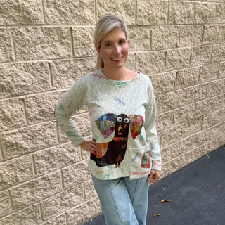 A woman wearing a relaxed ivory long-sleeve crew neck tunic with a multicolor patchwork dachshund graphic on the front and a functional hanging pocket, paired with wide-leg light-wash jeans.