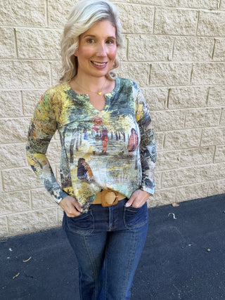 A woman wearing a long-sleeve v-slit burnout top in a multicolor scenic park print featuring dog walkers among autumn-toned trees in gold, green, and grey, paired with dark-wash jeans and a woven belt.