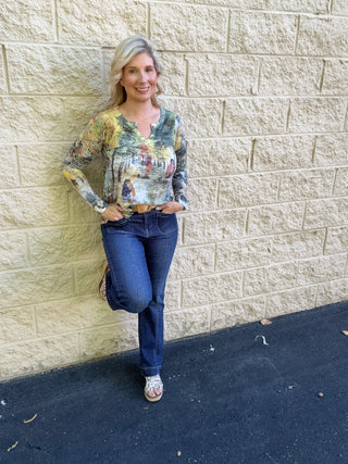 A woman wearing a long-sleeve v-slit burnout top in a multicolor scenic park print featuring dog walkers among autumn-toned trees in gold, green, and grey, paired with dark-wash jeans and a woven belt.