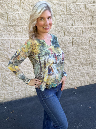 A woman wearing a long-sleeve v-slit burnout top in a multicolor scenic park print featuring dog walkers among autumn-toned trees in gold, green, and grey, paired with dark-wash jeans and a woven belt.