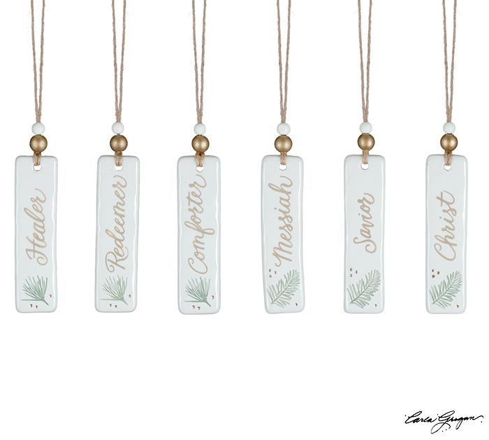 Six hand-painted ceramic tag ornaments with gold lettering and pine sprig accents reading Healer, Redeemer, Comforter, Messiah, Savior, and Christ — faith-inspired Christmas décor from The Pink Doxie.