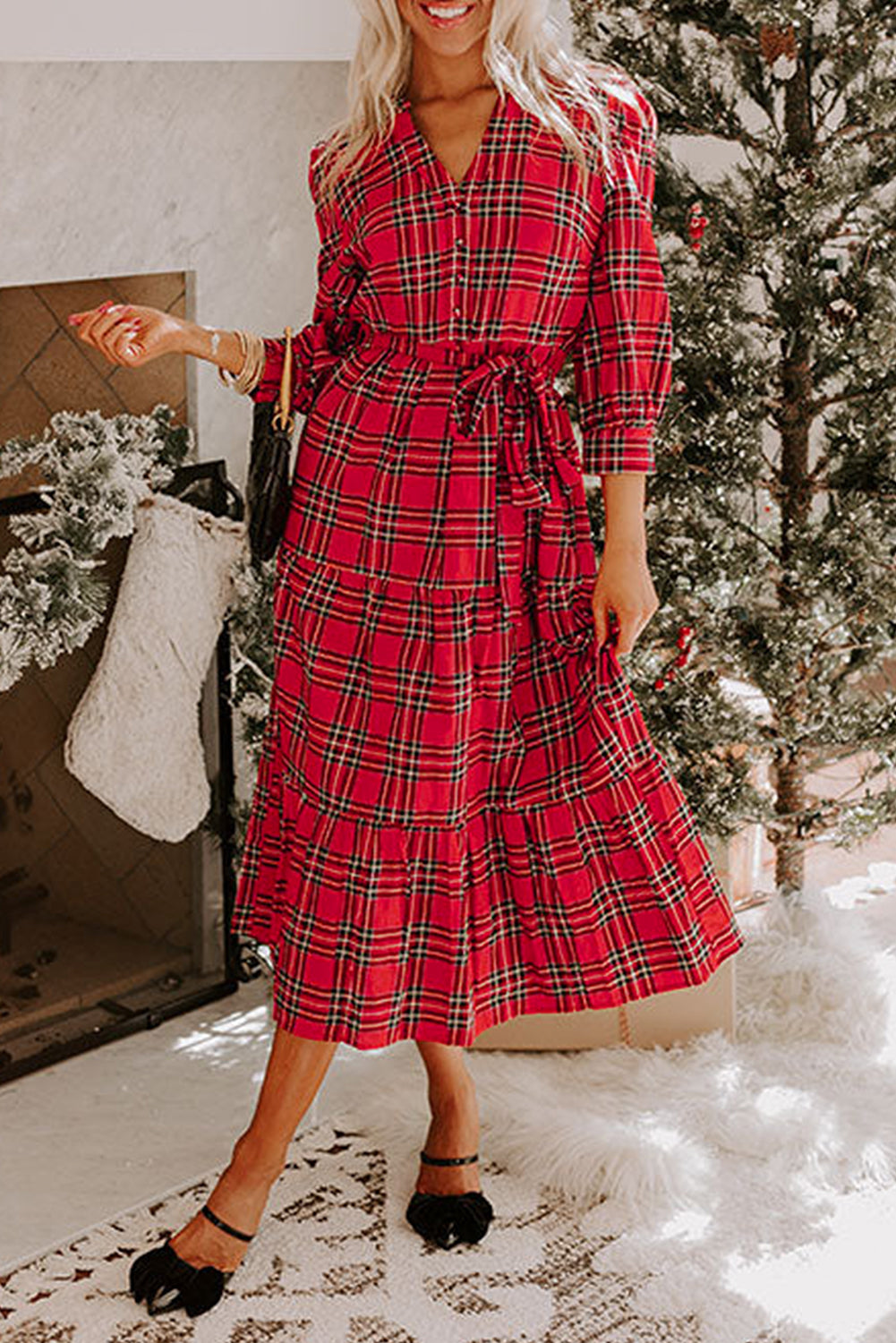 Woman wearing a red Christmas plaid long dress with a tie waist and ruffle hem, standing by a holiday fireplace and Christmas tree.